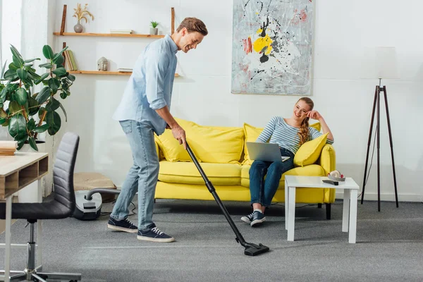 Homme souriant nettoyage tapis avec aspirateur près de petite amie en utilisant un ordinateur portable sur le canapé à la maison — Photo de stock