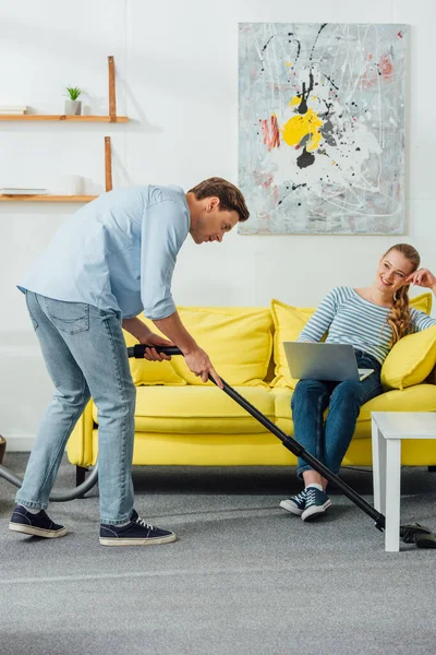 Bel homme nettoyage tapis dans le salon près de femme souriante avec ordinateur portable sur le canapé — Photo de stock