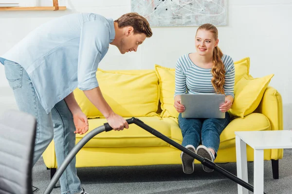 Selective focus of smiling woman with laptop looking at handsome boyfriend with vacuum cleaner in living room — Stock Photo