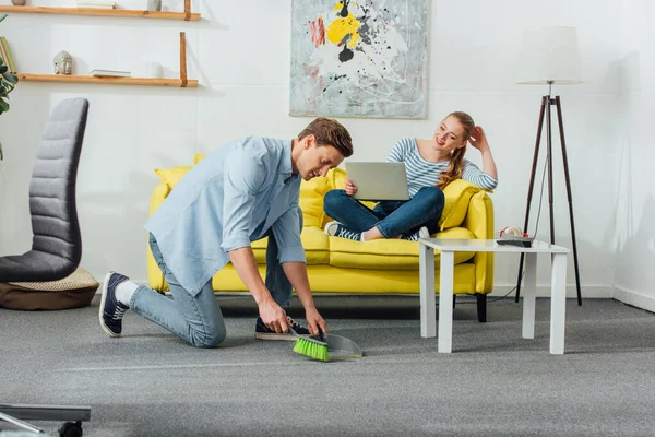 Man cleaning carpet with scoop and broom near smiling woman with laptop on couch — Stock Photo