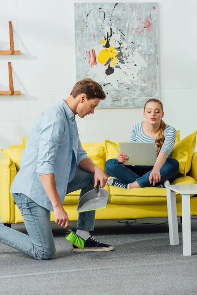 Woman with laptop pointing on carpet to man with scoop and broom in living room — Stock Photo