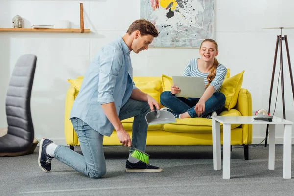 Smiling woman pointing on carpet to boyfriend with scoop and broom at home — Stock Photo