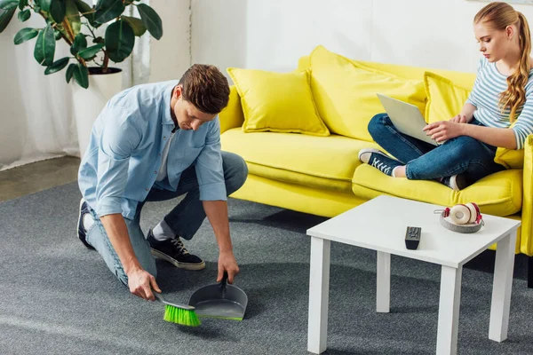 Hombre alfombra de limpieza con cuchara y escoba cerca de la novia utilizando el ordenador portátil en el sofá - foto de stock