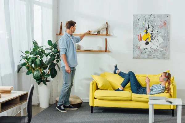 Seitenansicht einer Frau mit Kopfhörer per Smartphone auf der Couch und ihres Freundes, der zu Hause Regale mit Staubbürste putzt — Stockfoto
