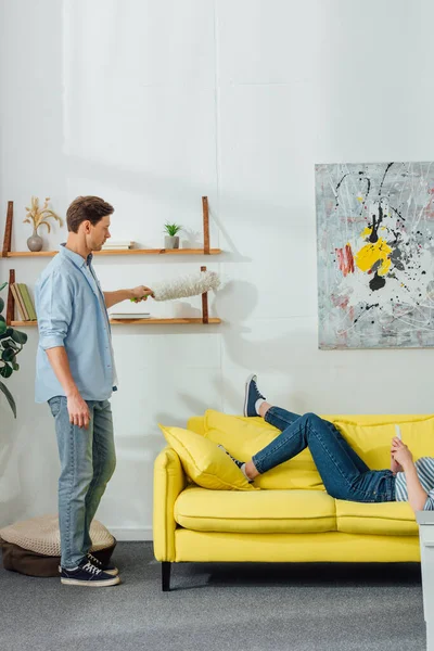 Side view of man cleaning shelves with dust brush near girlfriend using smartphone on sofa in living room — Stock Photo