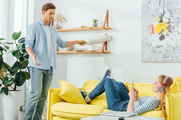 Man with dust brush looking at girlfriend in headphones using laptop on sofa at home — Stock Photo