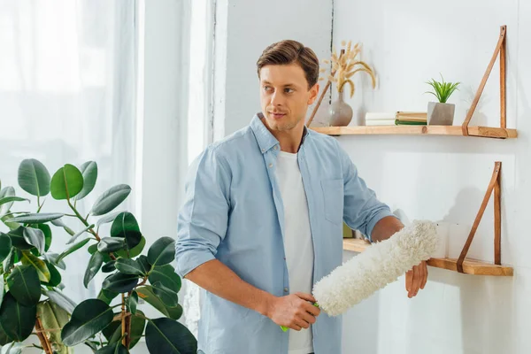 Handsome man holding dust brush near shelves with plants and books in living room — Stock Photo