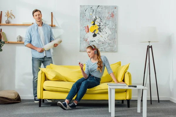 Hombre con cepillo de polvo sonriendo a la cámara cerca de su novia en los auriculares con el teléfono inteligente en el sofá en casa - foto de stock