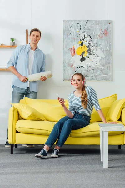 Smiling man with dust brush and woman in headphones with smartphone looking at camera in living room — Stock Photo