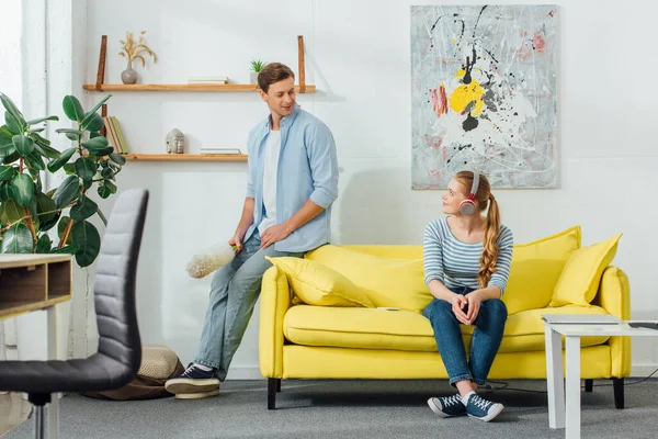 Man with dust brush smiling at girlfriend in headphones on couch in living room — Stock Photo