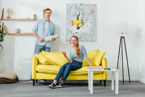 Mujer en auriculares con smartphone y novio con cepillo de polvo sonriendo a la cámara en la sala de estar - foto de stock