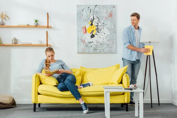 Homme avec chiffon nettoyage lampadaire près de petite amie lecture livre sur canapé dans le salon — Photo de stock