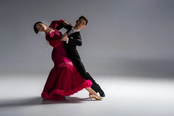 Élégant jeune couple de danseurs de salon en robe rouge en costume dansant sur gris — Photo de stock