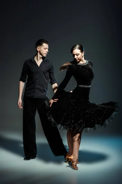 Elegante pareja joven de bailarines de salón en trajes negros bailando en la oscuridad - foto de stock