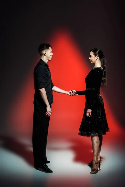 Élégant jeune couple de danseurs de salon en tenues noires dansant en lumière rouge — Photo de stock