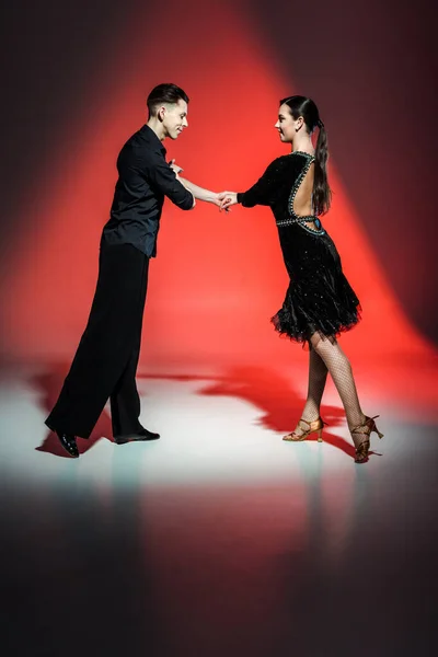 Elegante pareja joven de bailarines de salón en trajes negros bailando en luz roja - foto de stock