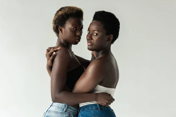 Афроамериканські жінки в джинсах і вершинах обіймаються ізольовано на сірому — стокове фото