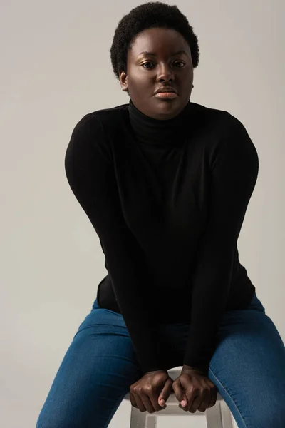 Giovane donna afroamericana in jeans e dolcevita nero seduta su sgabello isolato su grigio — Foto stock