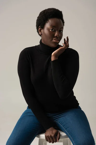 Beautiful african american girl in jeans and black turtleneck sitting on stool isolated on grey — Stock Photo