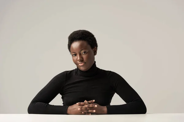 Beautiful african american woman in black turtleneck sitting at table isolated on grey — Stock Photo