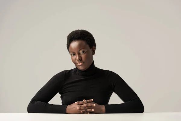 Hermosa mujer afroamericana en cuello alto negro sentada a la mesa aislada en gris - foto de stock