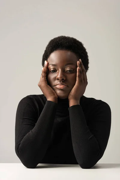 Femme afro-américaine bouleversée au col roulé noir assis à la table isolé sur gris — Photo de stock