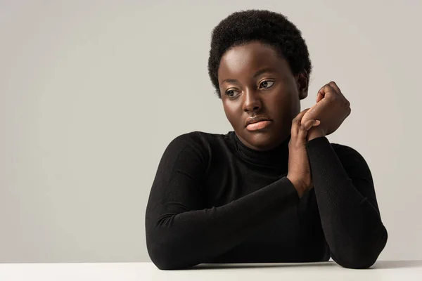 Mulher afro-americana pensiva em gola alta preta sentada à mesa isolada em cinza — Fotografia de Stock