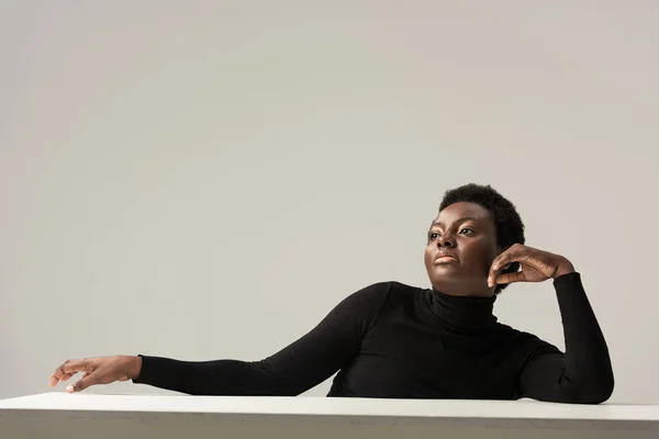 Fiduciosa donna africana americana in dolcevita nero seduta a tavola isolata sul grigio — Foto stock
