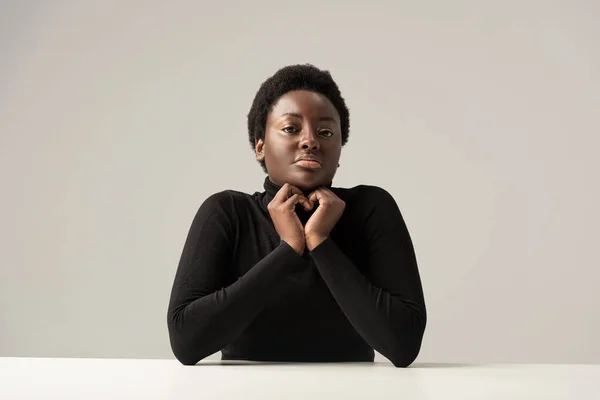 Belle femme afro-américaine au col roulé noir assise à table isolée sur gris — Photo de stock