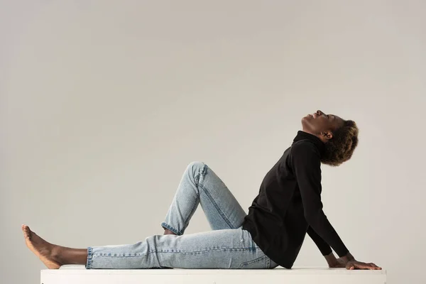 Heureuse afro-américaine fille en jeans et col roulé noir assis sur cube blanc isolé sur gris — Photo de stock