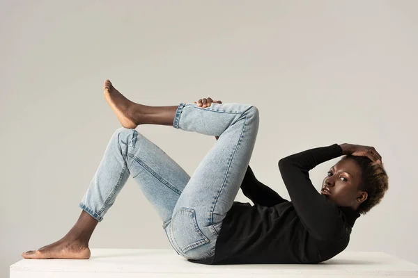 Африканская американка в джинсах и черной водолазке сидит на белом кубе изолированный на сером — стоковое фото