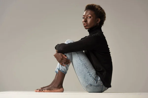 Joven afroamericana mujer en jeans y cuello alto negro sentado en cubo blanco aislado en gris - foto de stock