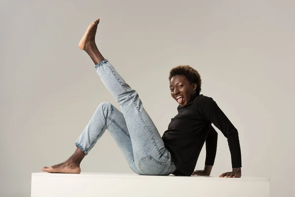 Funny african american girl in jeans and black turtleneck sticking tongue out while sitting on white cube isolated on grey — Stock Photo