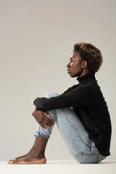 Barefoot african american girl in black turtleneck sitting on white cube isolated on grey — Stock Photo