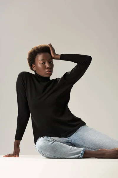 African american girl in black turtleneck sitting on white cube isolated on grey — Stock Photo