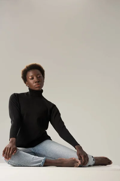 Beautiful african american girl in black turtleneck sitting on white cube isolated on grey — Stock Photo