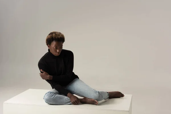 Africano americano ragazza in nero dolcevita seduta su bianco cubo su grigio — Foto stock