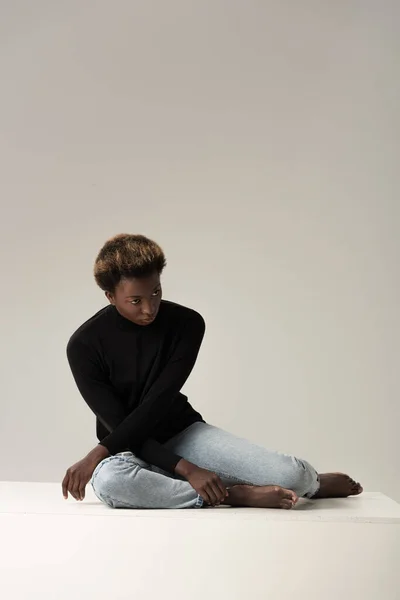 Chica afroamericana en cuello alto negro sentado en cubo blanco aislado en gris - foto de stock