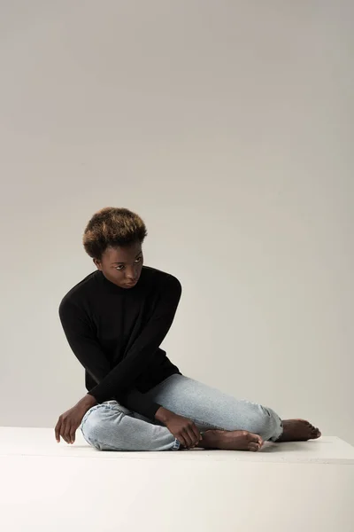 African american girl in black turtleneck sitting on white cube isolated on grey — Stock Photo
