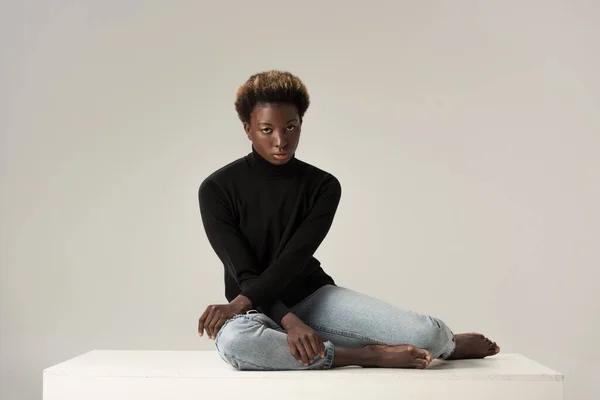 Attractive african american girl in jeans and black turtleneck sitting on white cube isolated on grey — Stock Photo