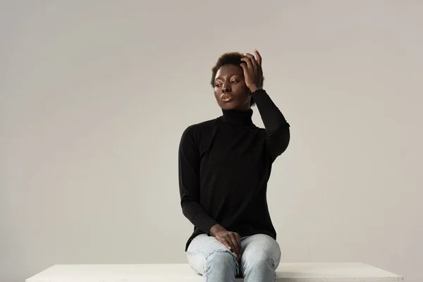 Menina afro-americana macia em jeans e gola alta preta sentado em cubo branco isolado em cinza — Fotografia de Stock