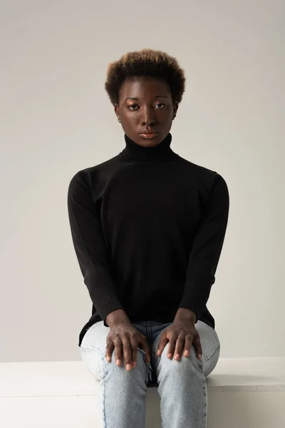 Afroamerikanerin in Jeans und schwarzem Rollkragen sitzt auf weißem Würfel isoliert auf grau — Stockfoto