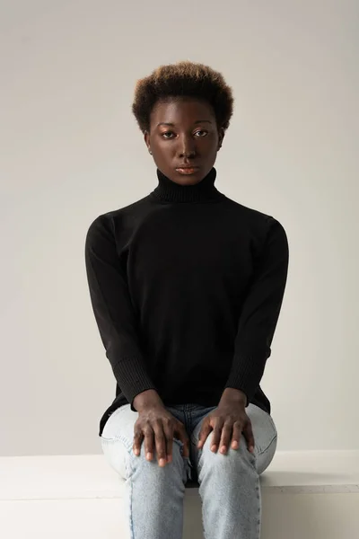 African american girl in jeans and black turtleneck sitting on white cube isolated on grey — Stock Photo