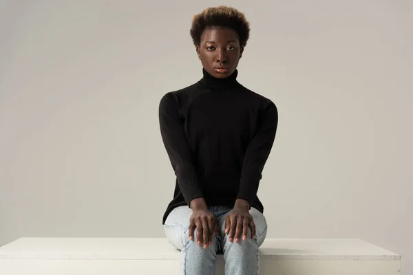 Beautiful african american girl in jeans and black turtleneck sitting on white cube isolated on grey — Stock Photo