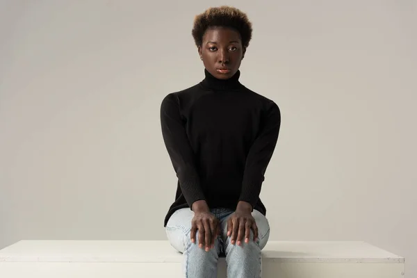 Hermosa chica afroamericana en jeans y cuello alto negro sentado en cubo blanco aislado en gris - foto de stock