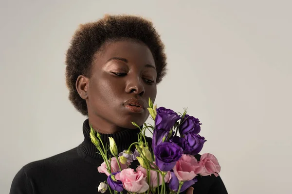 Hermosa mujer afroamericana en cuello alto negro sosteniendo flores de eustoma aisladas en gris - foto de stock