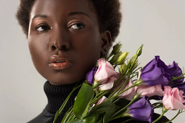 Mujer afroamericana en cuello alto negro sosteniendo flores de eustoma aisladas en gris - foto de stock
