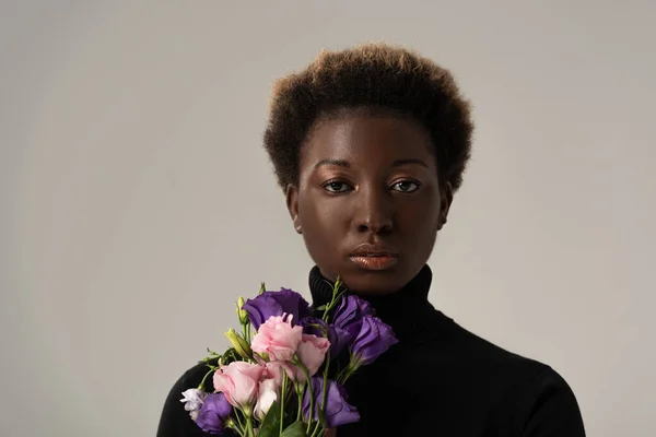 Приваблива афро-американська жінка в чорному черепашці тримає квіти еустоми ізольовані на сірому — стокове фото