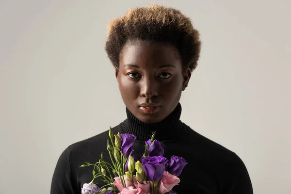 Afrikanerin mit schwarzem Rollkragen hält Eustoma-Blüten isoliert auf grau — Stockfoto