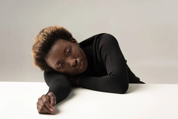 Femme afro-américaine fatiguée à col roulé noir assis sur la table isolé sur gris — Photo de stock